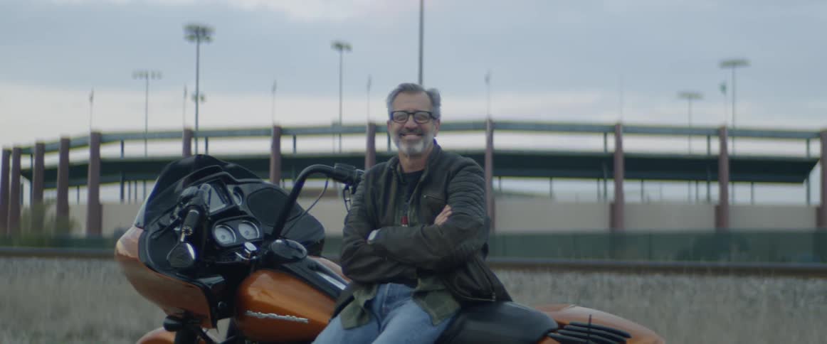Man wearing glasses smiles as he leans against a motorcycle
