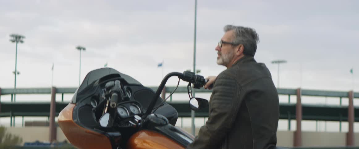 A man sits on a parked motorcycle and looks up at the sky