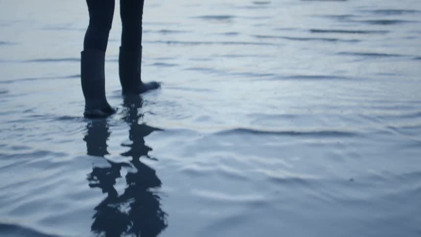 A woman wearing boots wades into the surf at sunset