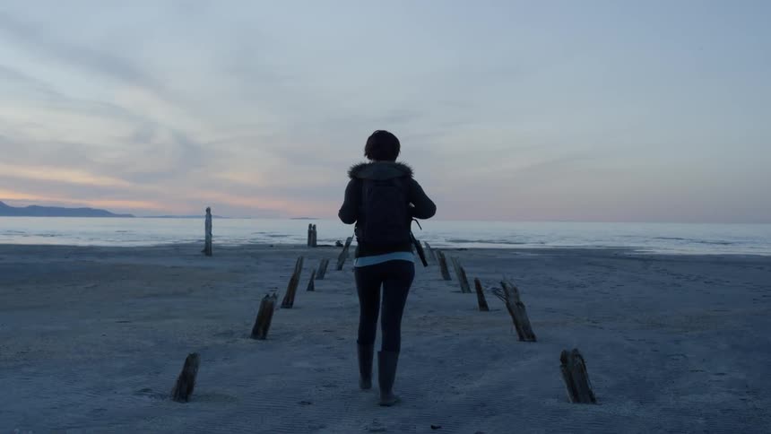 Woman wearing a jacket walks on the sand towards the lake