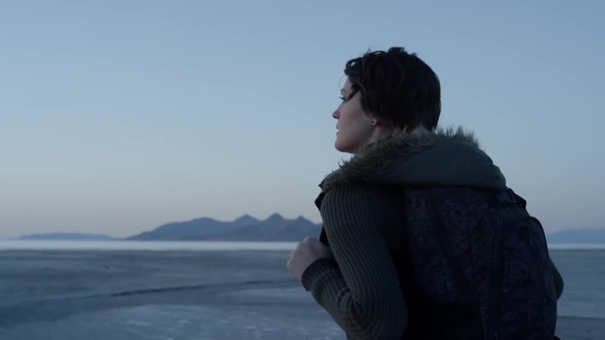 A woman wearing a backpack walks toward the ocean at sunset