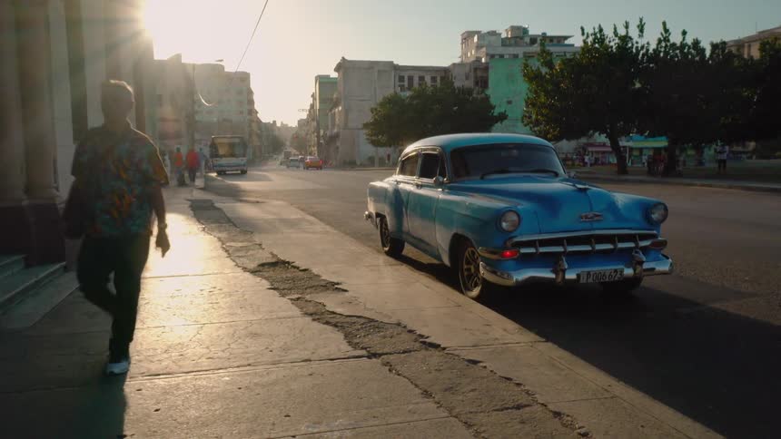Old blue car sits parked next to a sidewalk as a man walks by