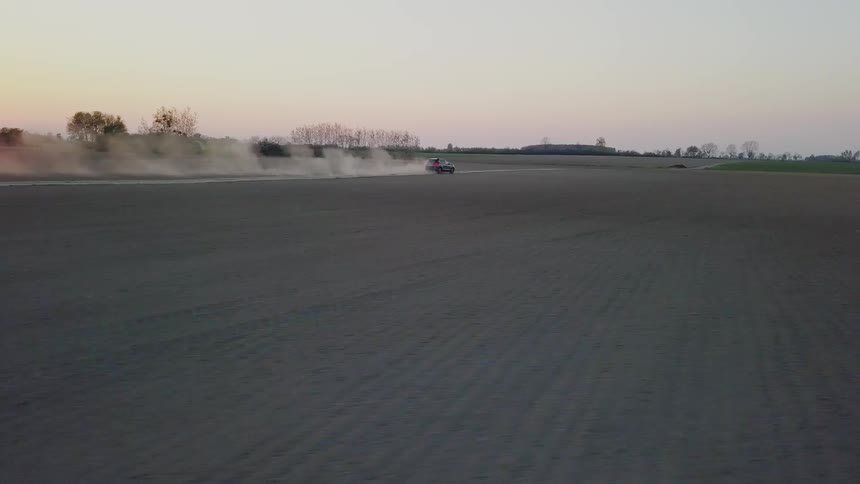 A vehicle is kicking up dust as it drives across a dirt road in a rural area at sunset