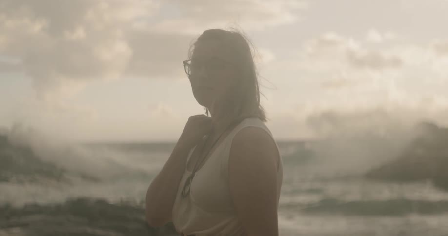 Woman wearing glasses stands near the ocean under a cloudy sky
