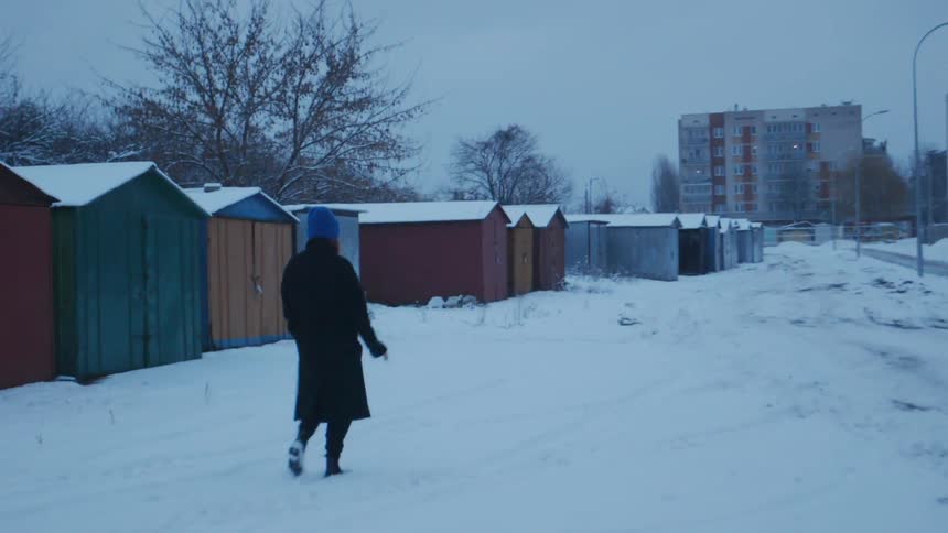 A woman smokes as she walks through the snow passing sheds