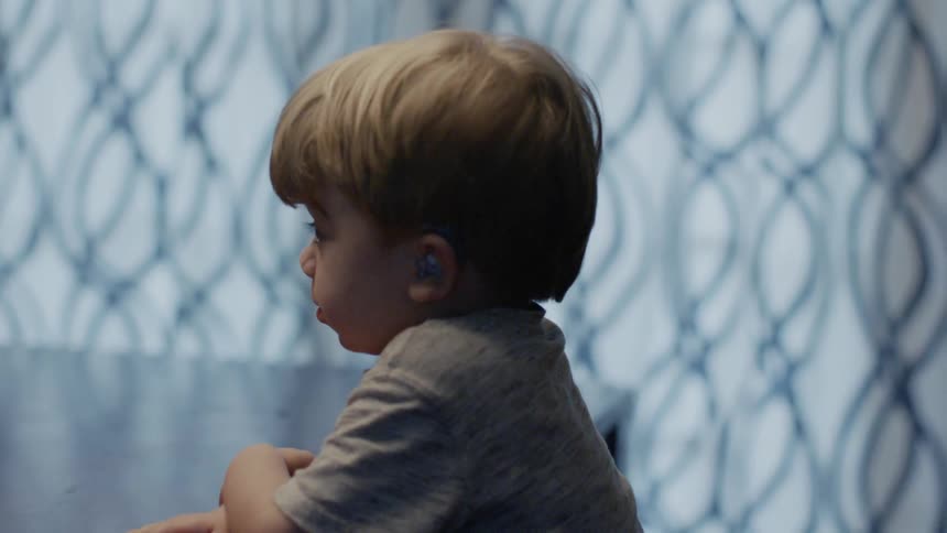 A young child wearing a hearing aid sits at a table