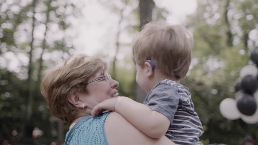 An older woman is kissing and carrying a toddler boy outside in daylight