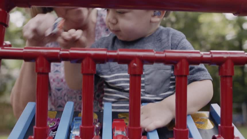 A woman holds a boy and helps him load toy cars on a track