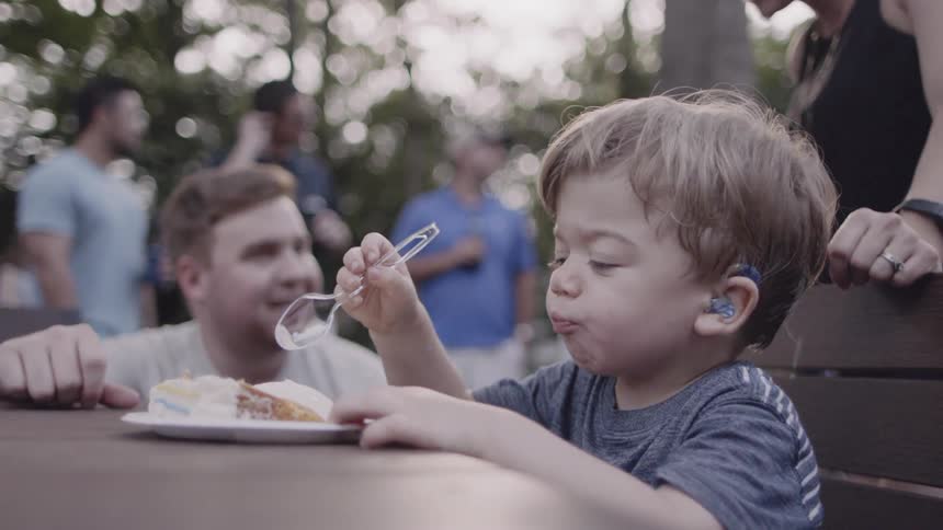 Toddler eats cake with a plastic spoon as a man watches