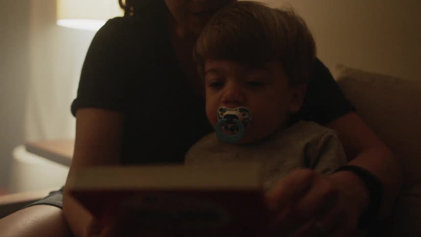 A woman is holding a toddler on her lap and opening a book as they sit inside in daylight