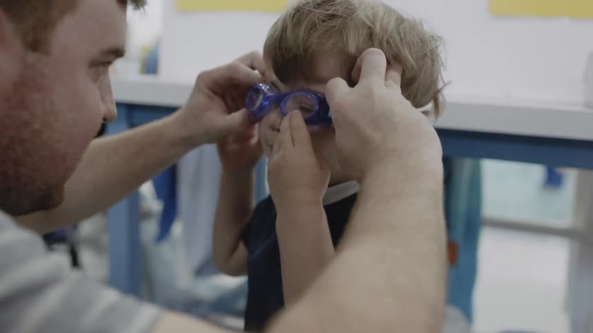 A man is helping a toddler boy put on swimming goggles