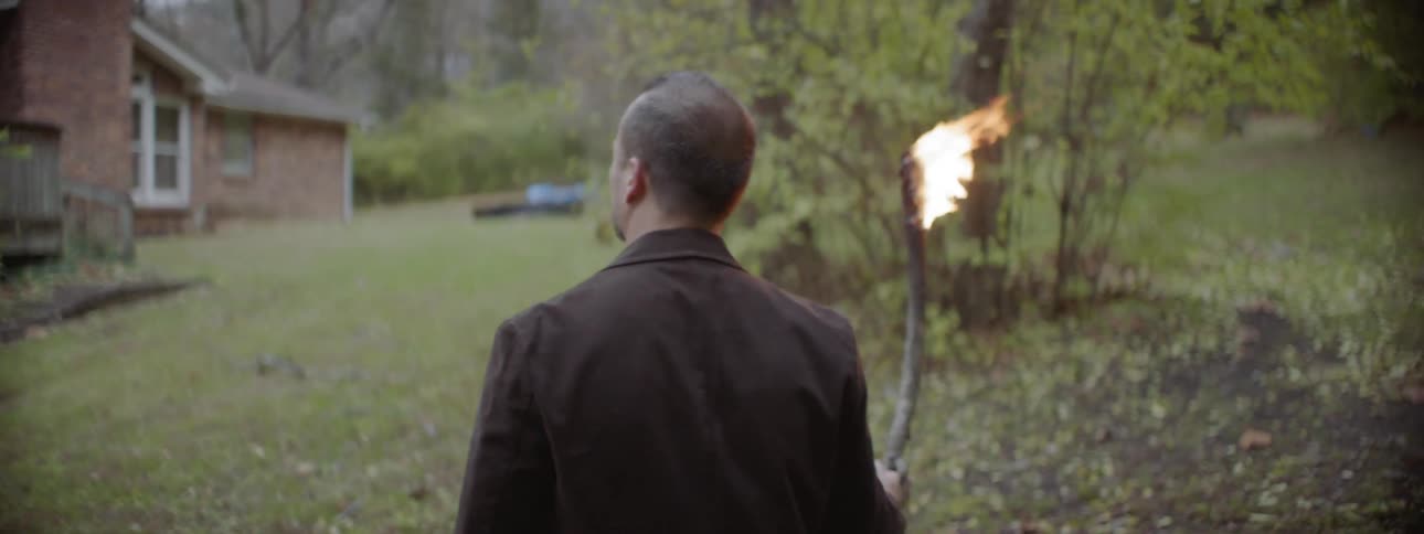 Man holding a burning stick of fire in a field near a house