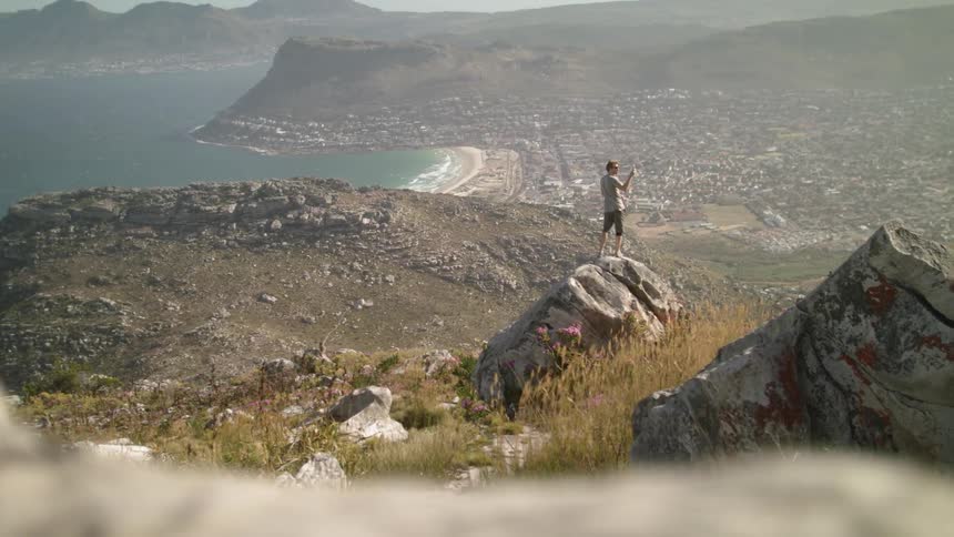 A woman is standing on a boulder overlooking a coastal community on a sunny day