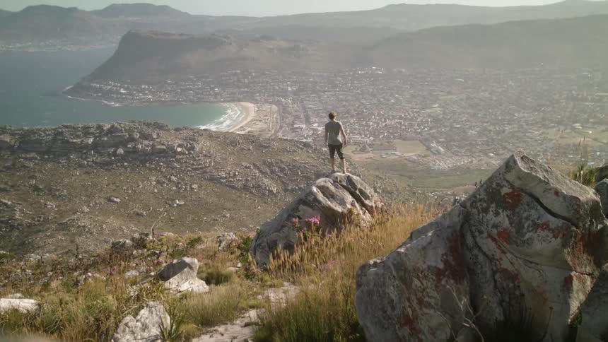 Man stands on a large rock on hilly landscape overlooking a body of water