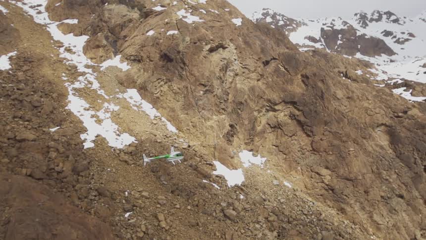 Helicopter travels close to the side of a rocky snow covered mountain