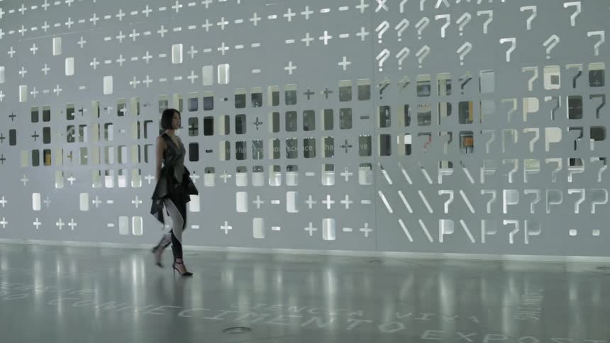 Woman walks near a wall with holes in it and light shining through