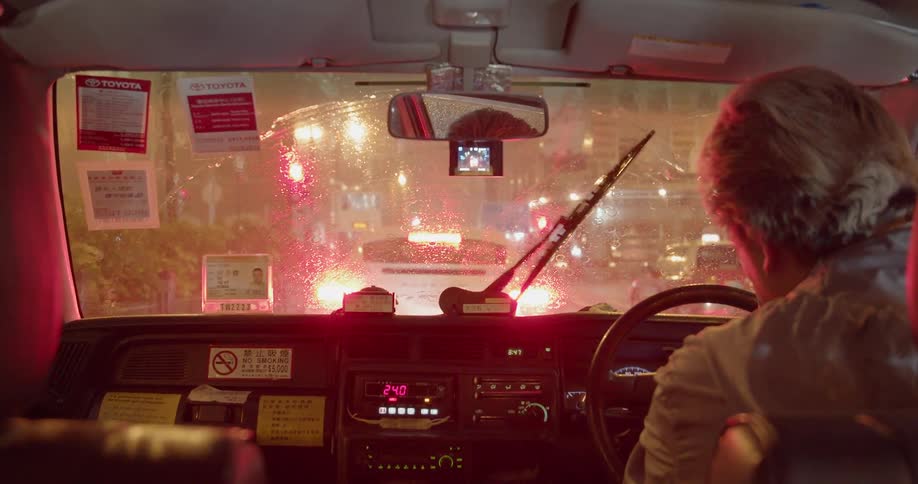 A person sits in city traffic on a rainy night with the windshield wipers going