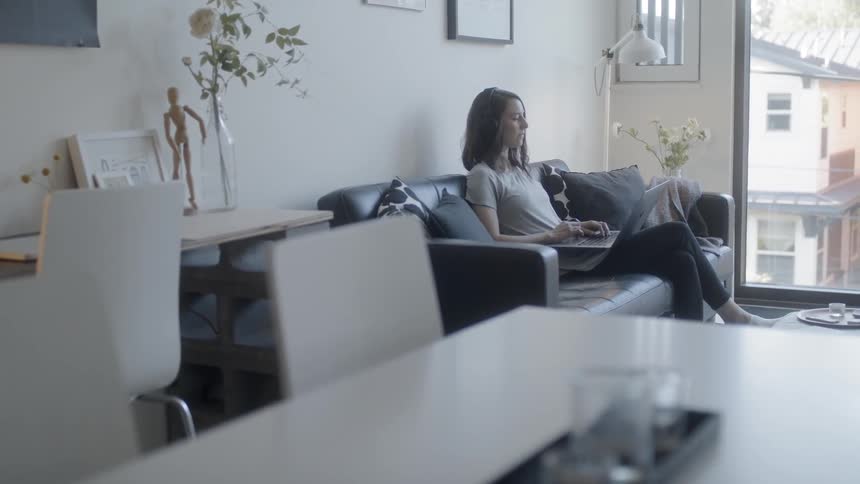 A woman is sitting on a couch typing on a laptop during the day
