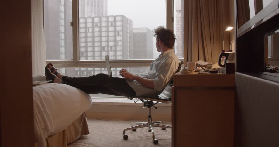 Man sits on a chair with legs on a bed typing on a laptop
