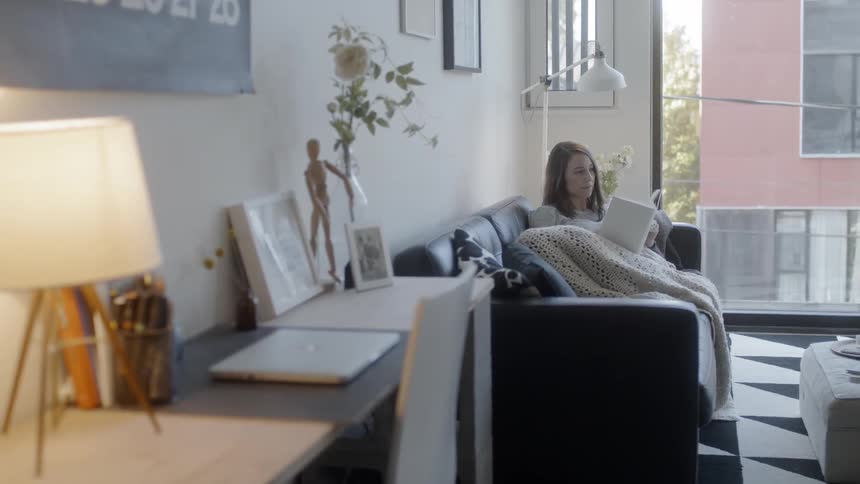 Woman sits on a couch with a blanket on her lap reading a book