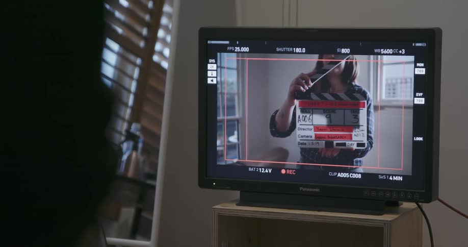 A person watches a monitor as a woman uses a clapperboard to indicate the start of a scene