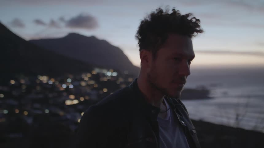 A man stands on a hilltop overlooking a town and the ocean at sunset