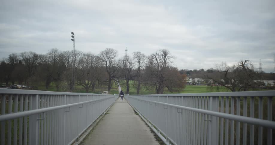 Woman jogs on a bridge toward trees on a cloudy day