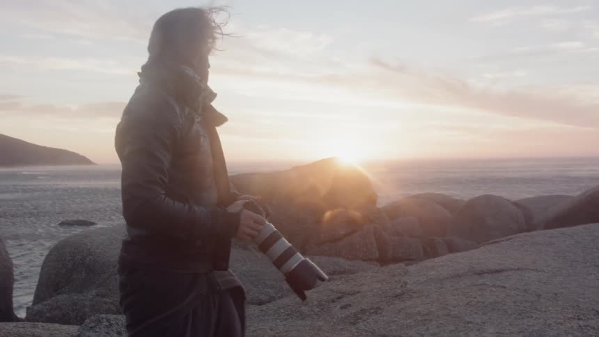 Man pushes hood off his head and looks down at his camera on a windy day near the water