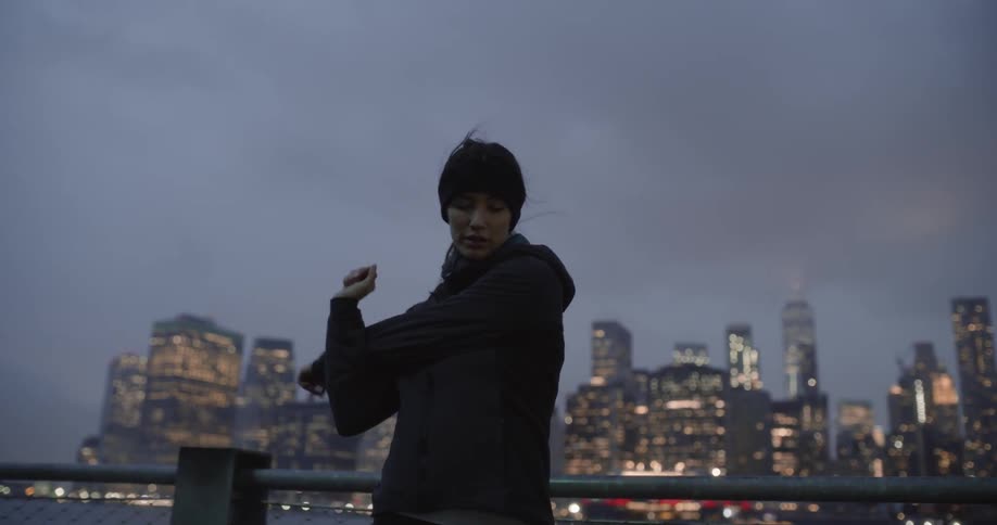 Woman stretches her arms next to a river with a city in the background