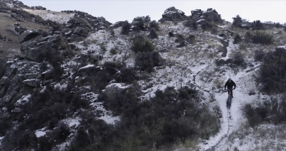 A young man riding his bike down a narrow, steep path on a rocky hillside that is dusted with snow