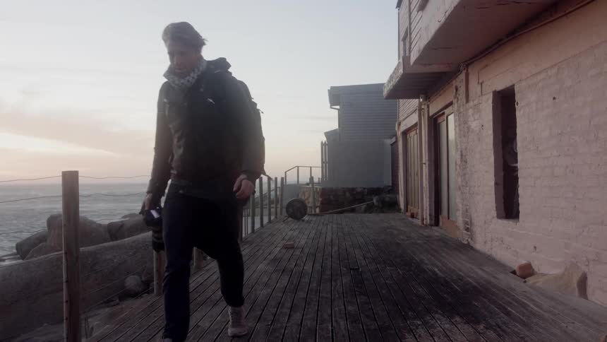 A man carrying a camera walks on a wooden boardwalk by the ocean