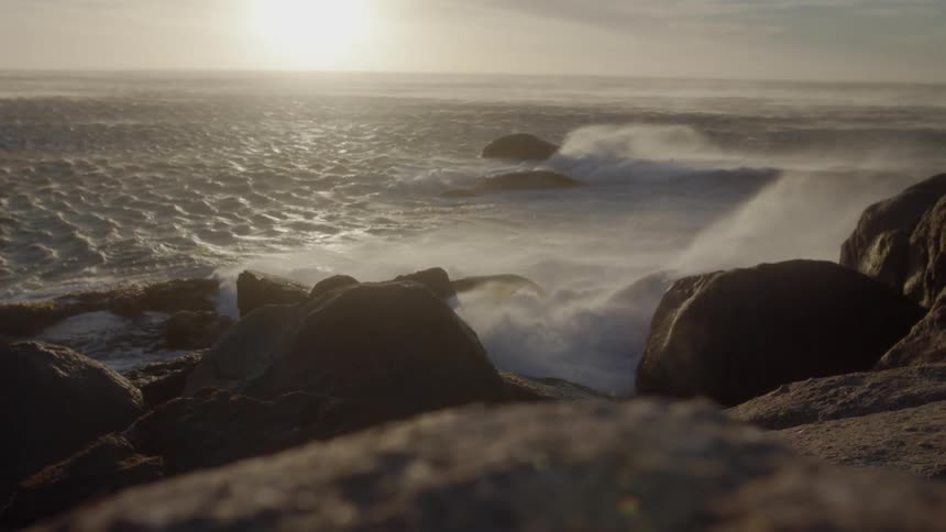 The wind is blowing mist off of waves as they crash over rocks in daylight
