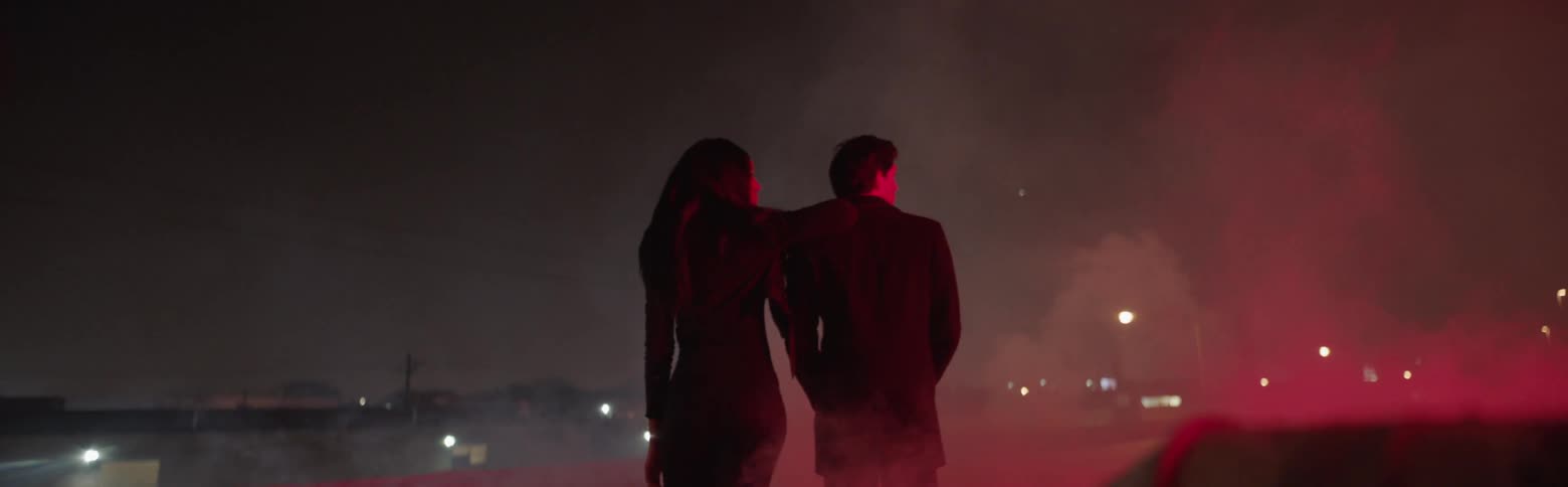 A woman is leaning on a man as they stand outside in a smokey setting at night