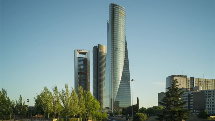 Traffic travels past skyscrapers as the wind blows trees on a sunny day