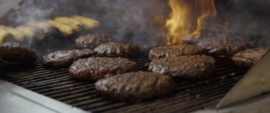 A man tends to burgers and corn cooking on a flaming grill