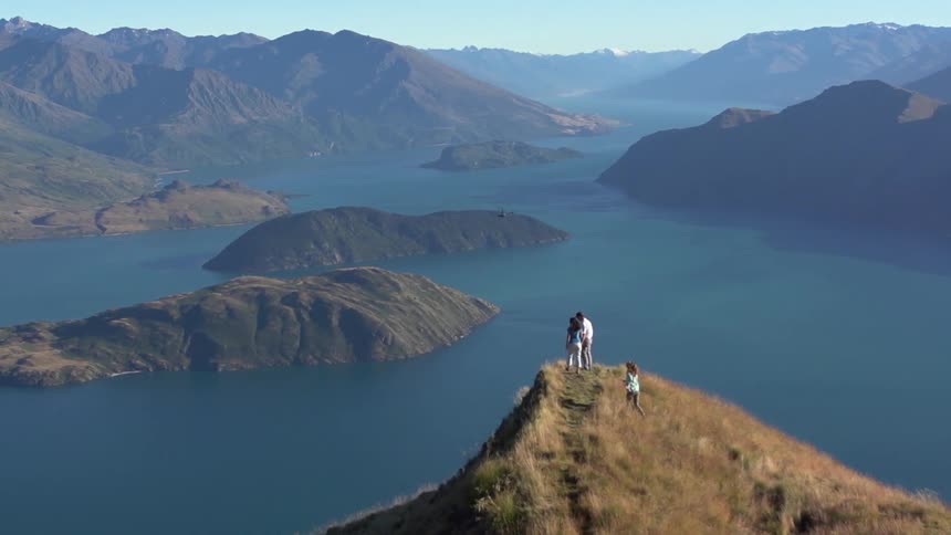 People standing on top of a cliff next to water located in a mountainous area while a helicopter approaches