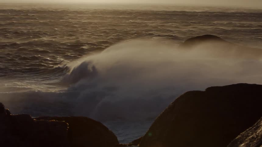 The wind is blowing mist off of waves as they roll toward rocks in daylight