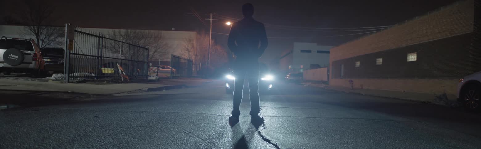 A man stands in front of a car with its headlights on
