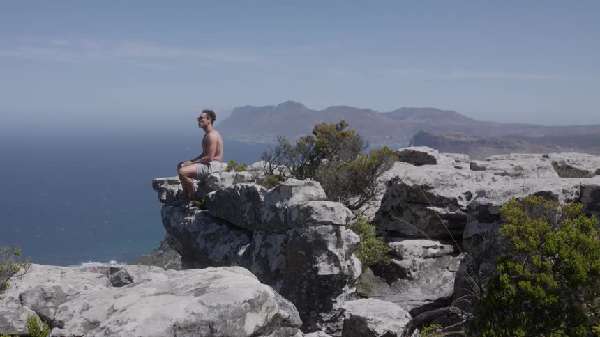 Man sits on a large rock on the edge of a cliff on a sunny day