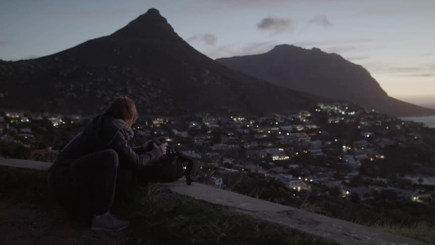 A man with a camera leans on a wall overlooking a town at sunset
