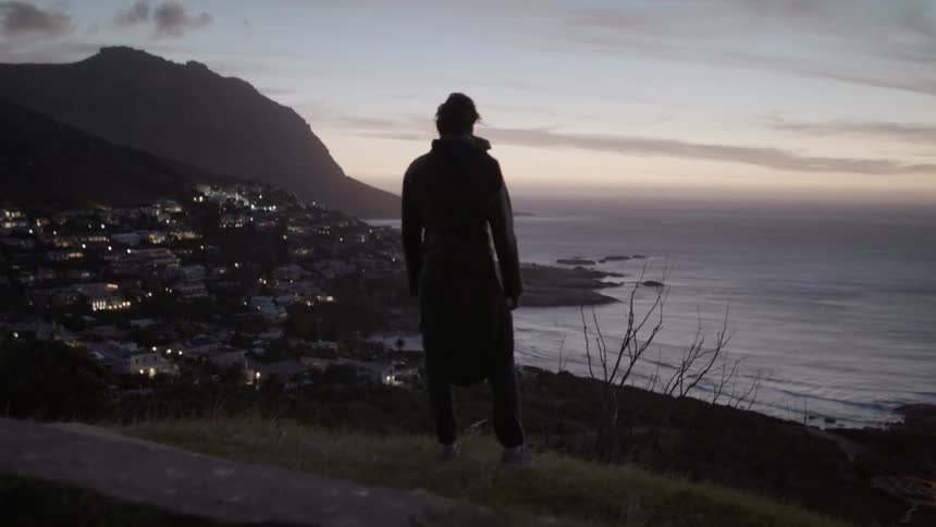 The person stands on the hill overlooking the shore as the sun sets