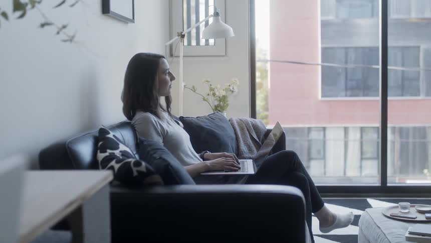 A woman sits on the couch typing on her laptop