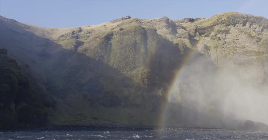 A rainbow sits over the water near the rocky hill