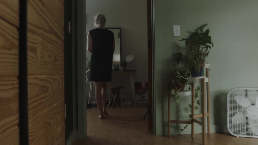 Woman walks in a hallway of a house and into a bedroom where a man sleeps