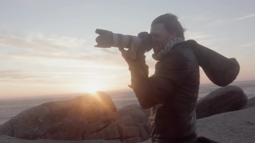 Man stands on a windy beach taking pictures at sunset