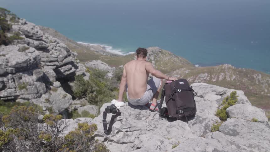 Man sits down on a rocky cliff holding a black backpack