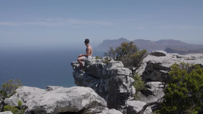 Man sits at the edge of a rocky cliff overlooking water