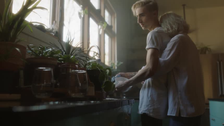 A woman hugs a man while he washes dishes at an overflowing sink
