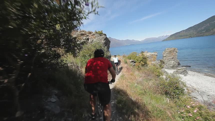 People riding mountain bikes along a narrow path located on a cliff next to a body of water surrounded by mountains