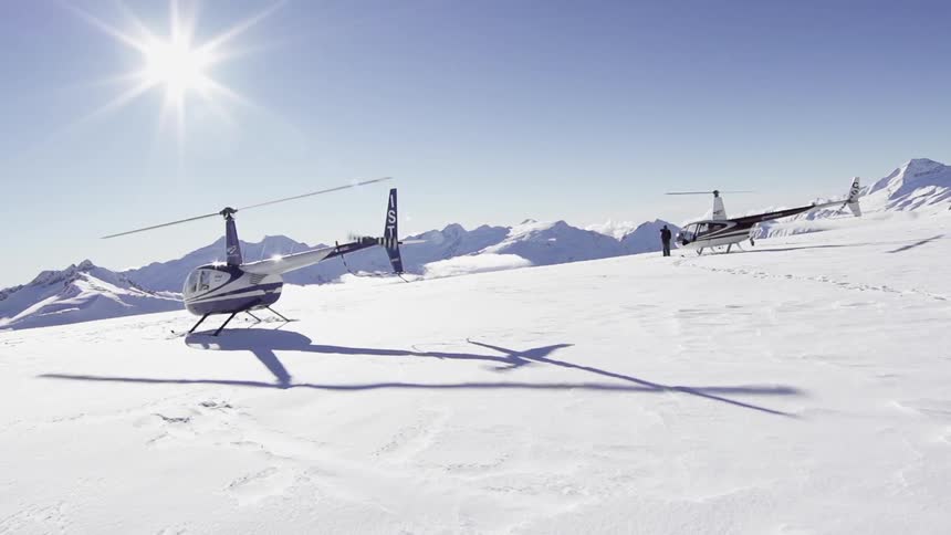 Two helicopters, with blades turning, resting on a snow covered mountain as some people stand or walk around one of the helicopters